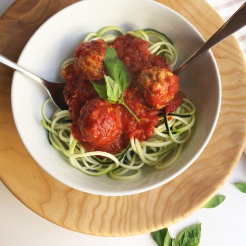 From above, on a wooden charger, a white bowl of zoodles and turkey meatballs with a silver fork and spoon, garnished with fresh basil.