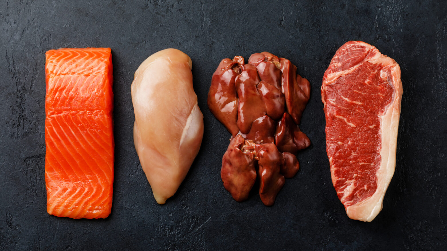 Raw cuts of salmon, chicken, steak, and organ meat on black slate
