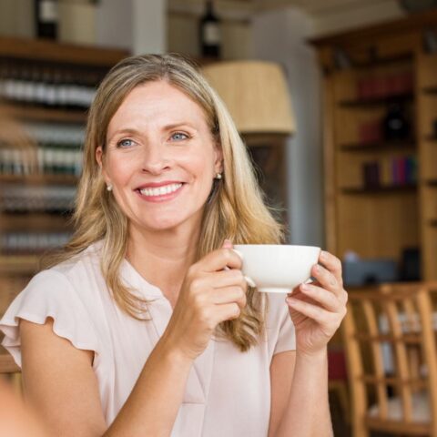 Beautiful blond woman smiles at a friend while enjoying tea in a warm room.
