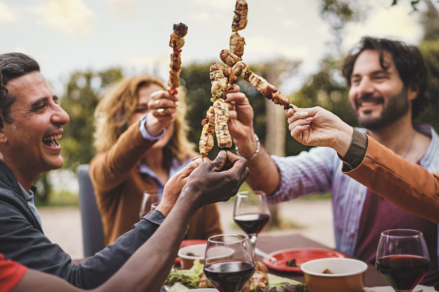 A group of friends having an outdoor dinner toast with skewers of meat.