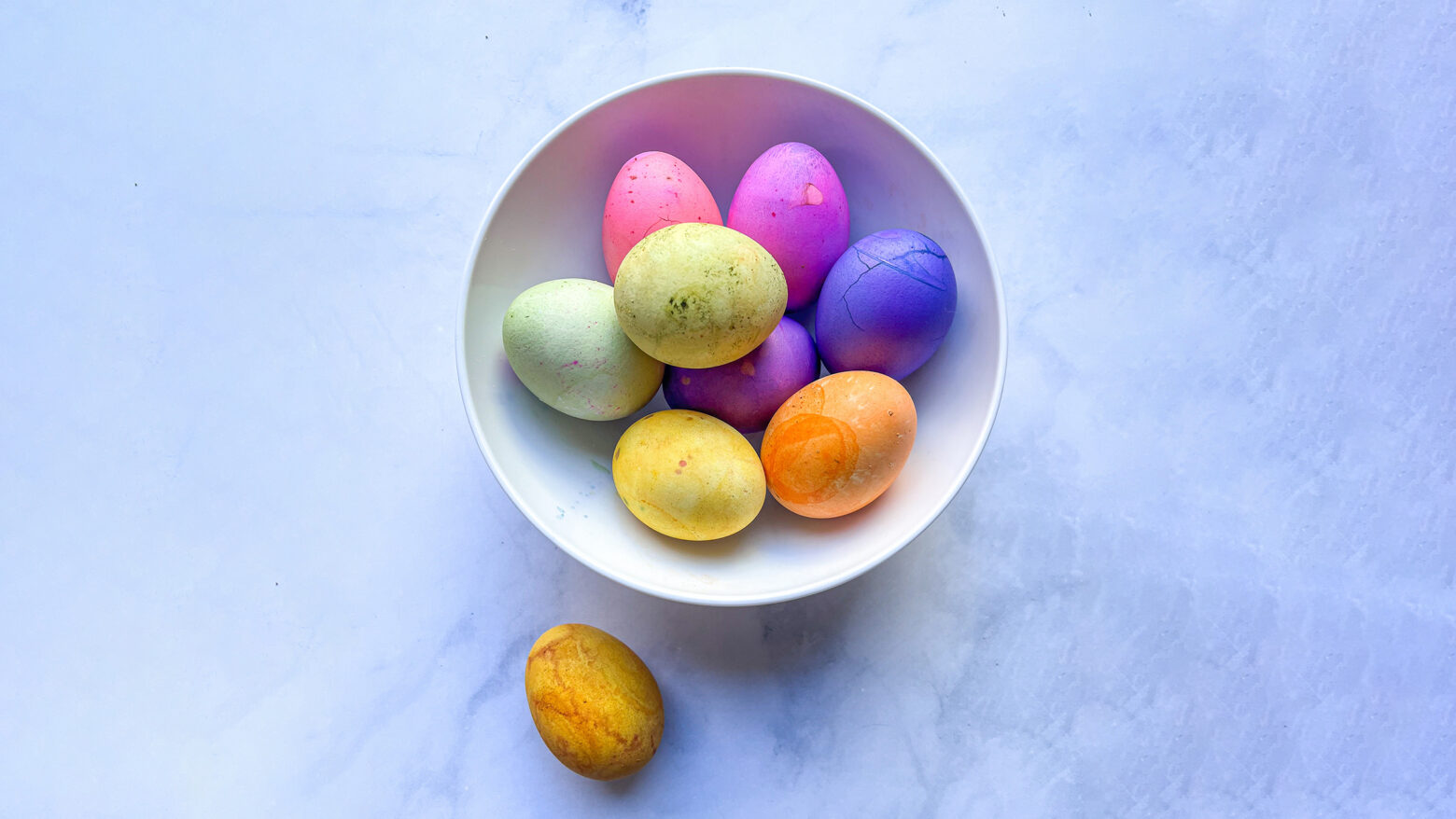 Colorful Easter eggs dyed with natural dyes sit in a white ceramic bowl on a white marble countertop.