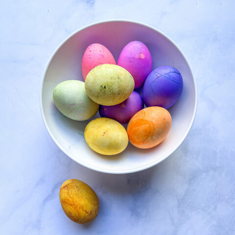 Colorful Easter eggs dyed with natural dyes sit in a white ceramic bowl on a white marble countertop.