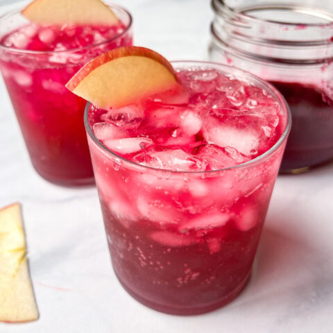 Red drink in glass with apple slice garnish.