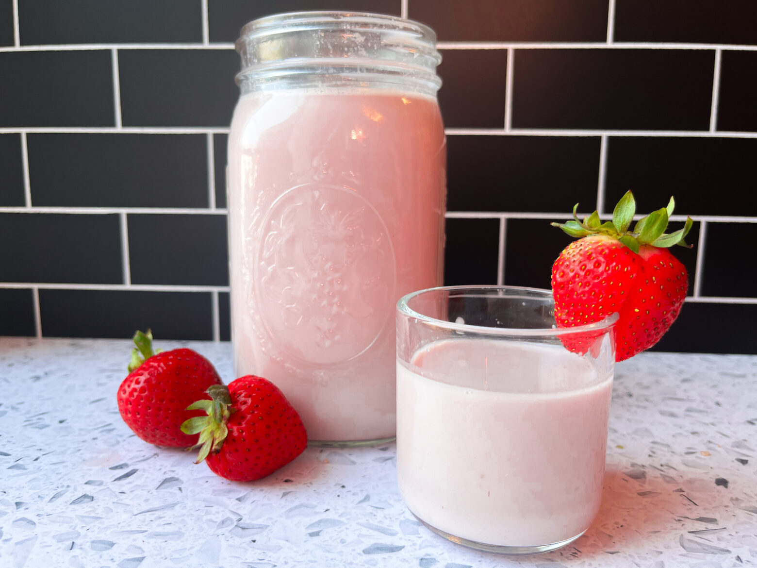 A clear glass container of strawberry almond milk next to another glass of milk