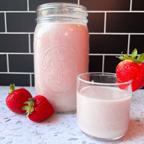 A clear glass container of strawberry almond milk next to another glass of milk