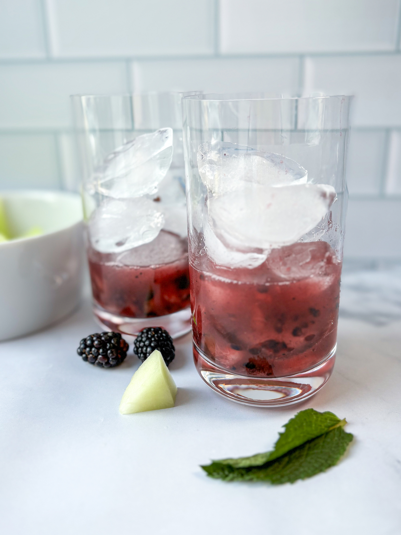 Straight on portrait view of two glasses filled with ice and partly filled with cocktail and mashed fruit, Garnishes of blackberries, mint leaf, and honeydew, Melon bowl in background