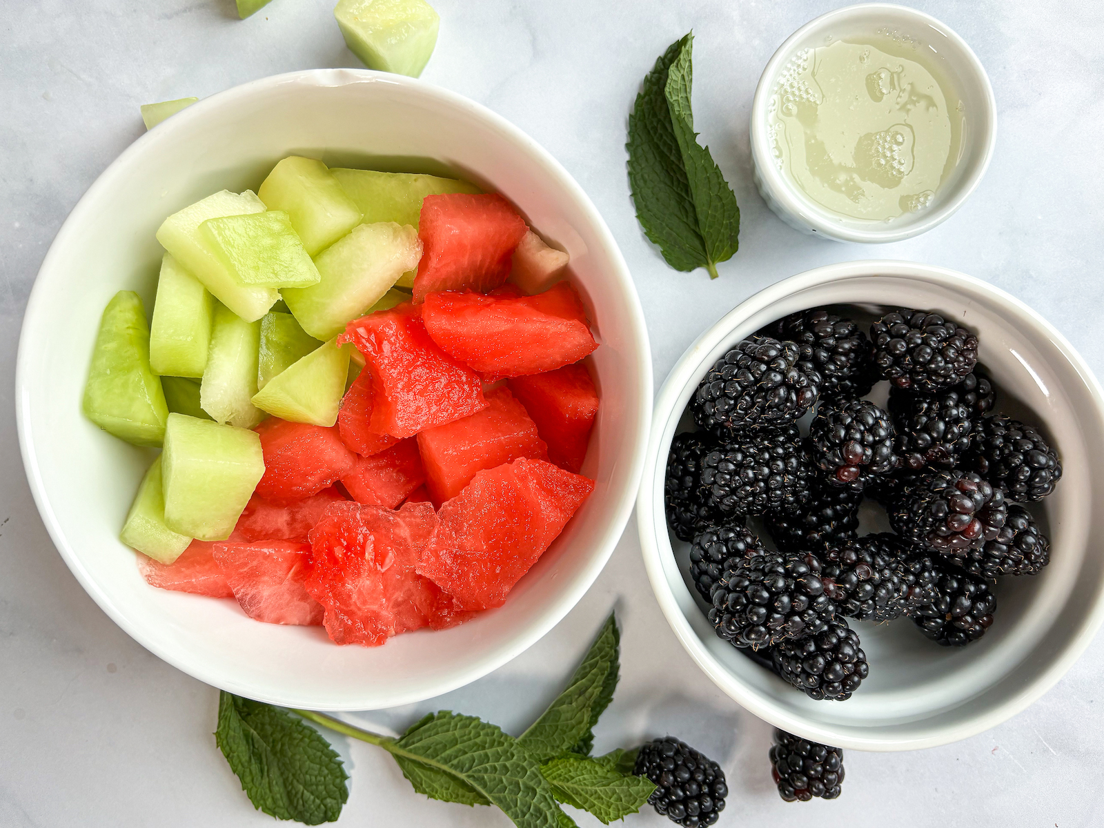 Birds eye horizontal view ingredients photo of honeydew, watermelon, blackberries and lychee juice, garnishes underneath