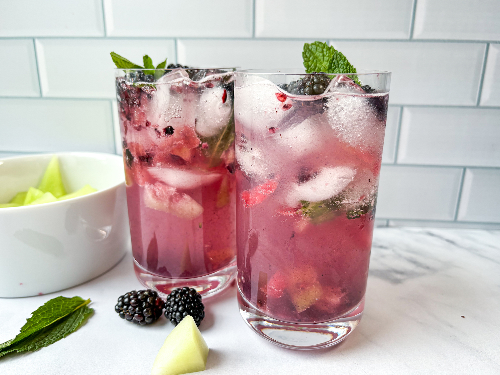 Horizontal straight on view of two finished cocktails with blackberry, melon and mint leaf garnishes, melon bowl in back