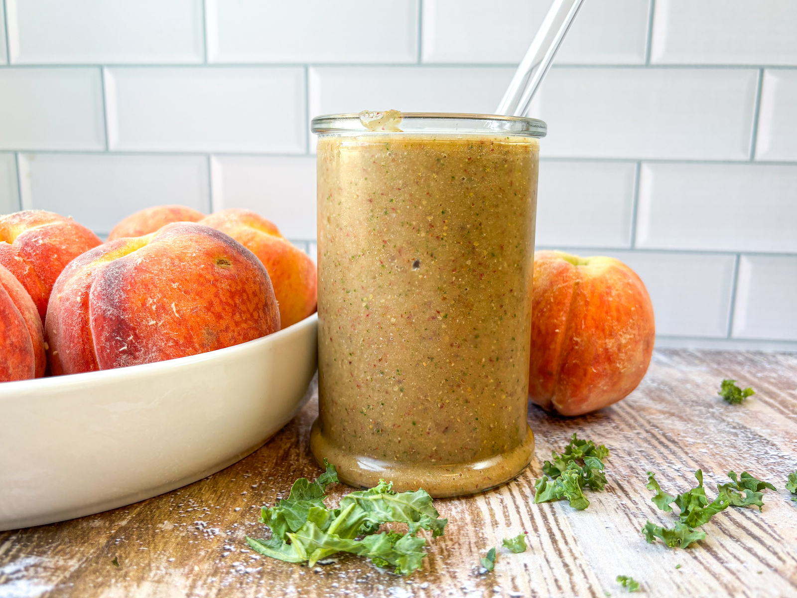 Straight on shot of finished smoothie in a glass. Kale garnish in front with peaches in a bowl and on table in the background