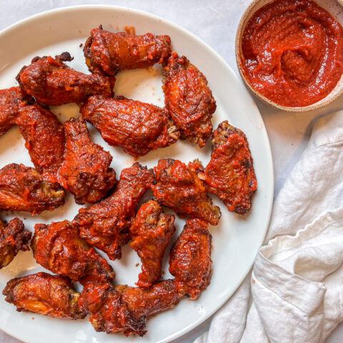 Birdseye view of finished Buffalo Beet BBQ Sauce Wings on a full plate with a dish of extra sauce and a towel next to the plate