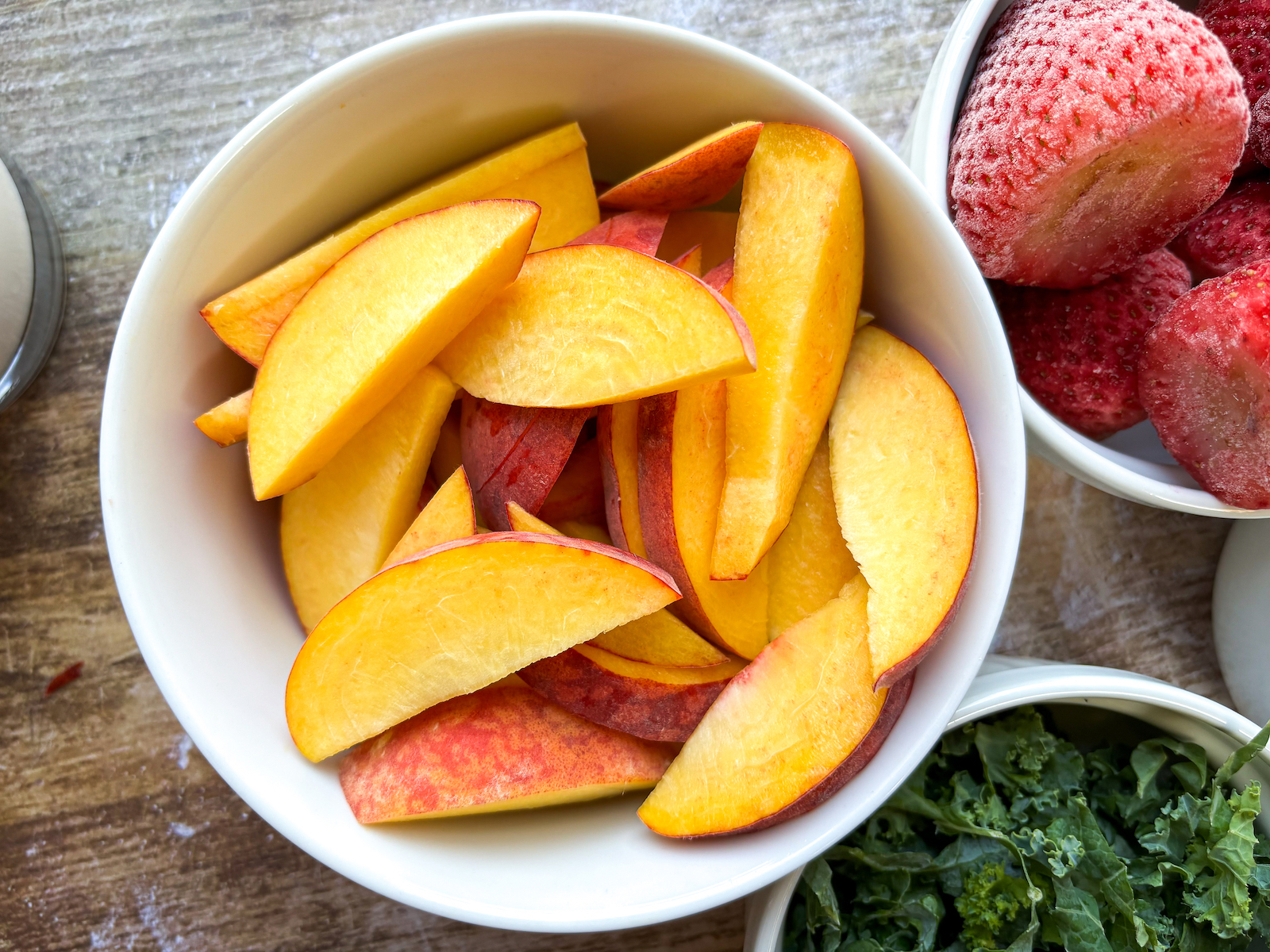 Peaches sliced in bowl from a bird eye view with kale and strawberries in the background