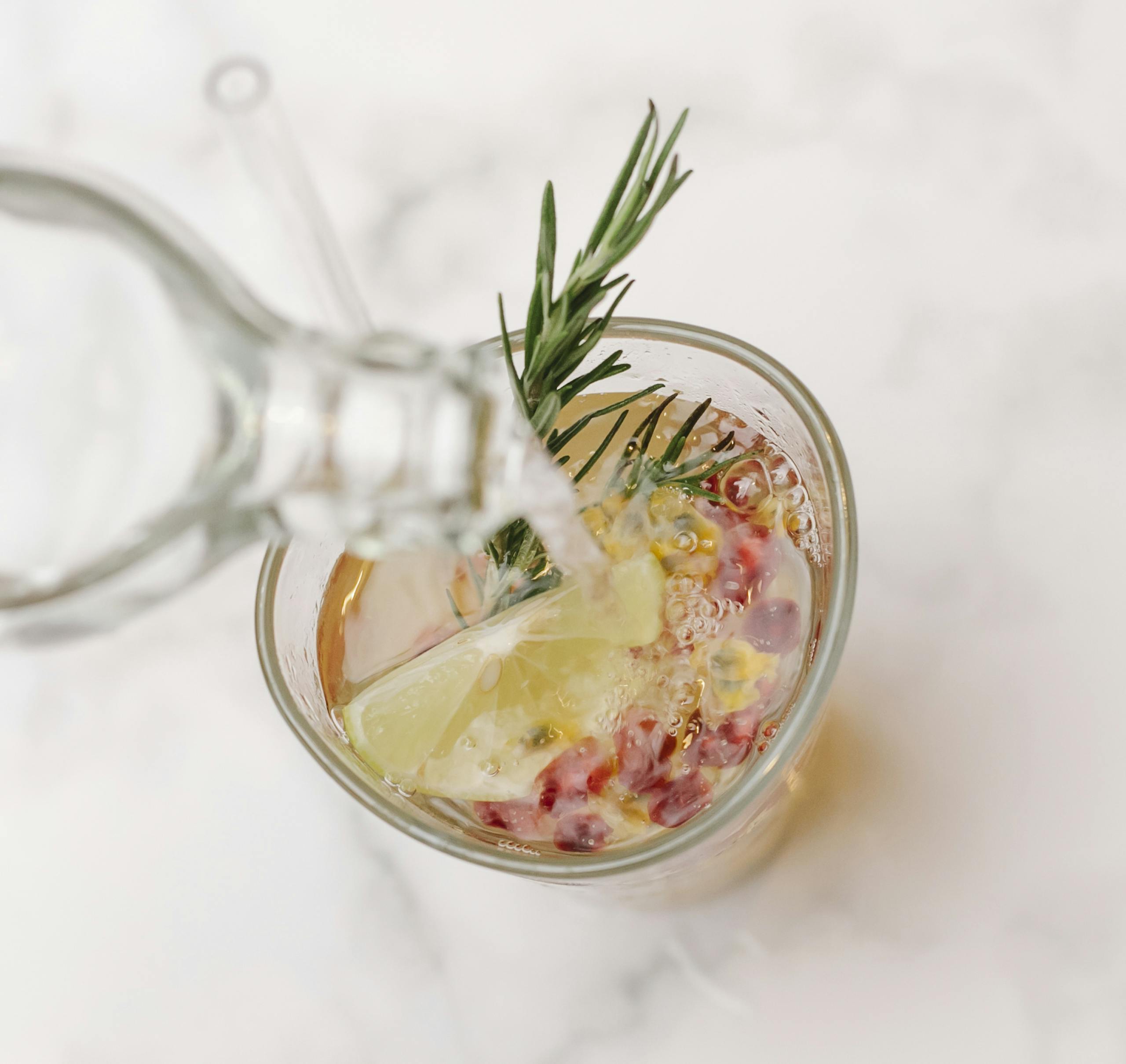 From above of glass bottle pouring water into glass with sprig of rosemary and slice of lemon placed on marble surface