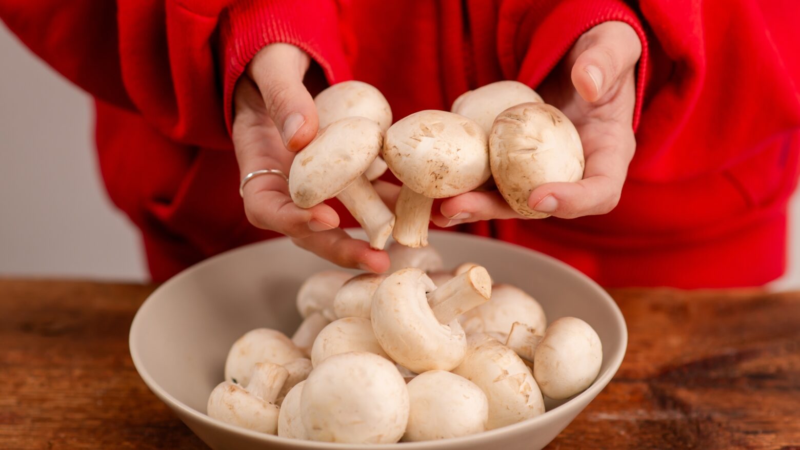 Close up of a woman in a red sweater holding white button mushrooms over a bowl of more mushrooms