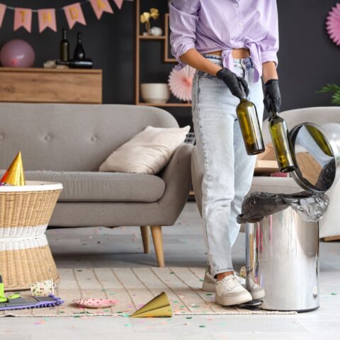 A woman throws empty wine bottles into a trash can while cleaning up after a party.