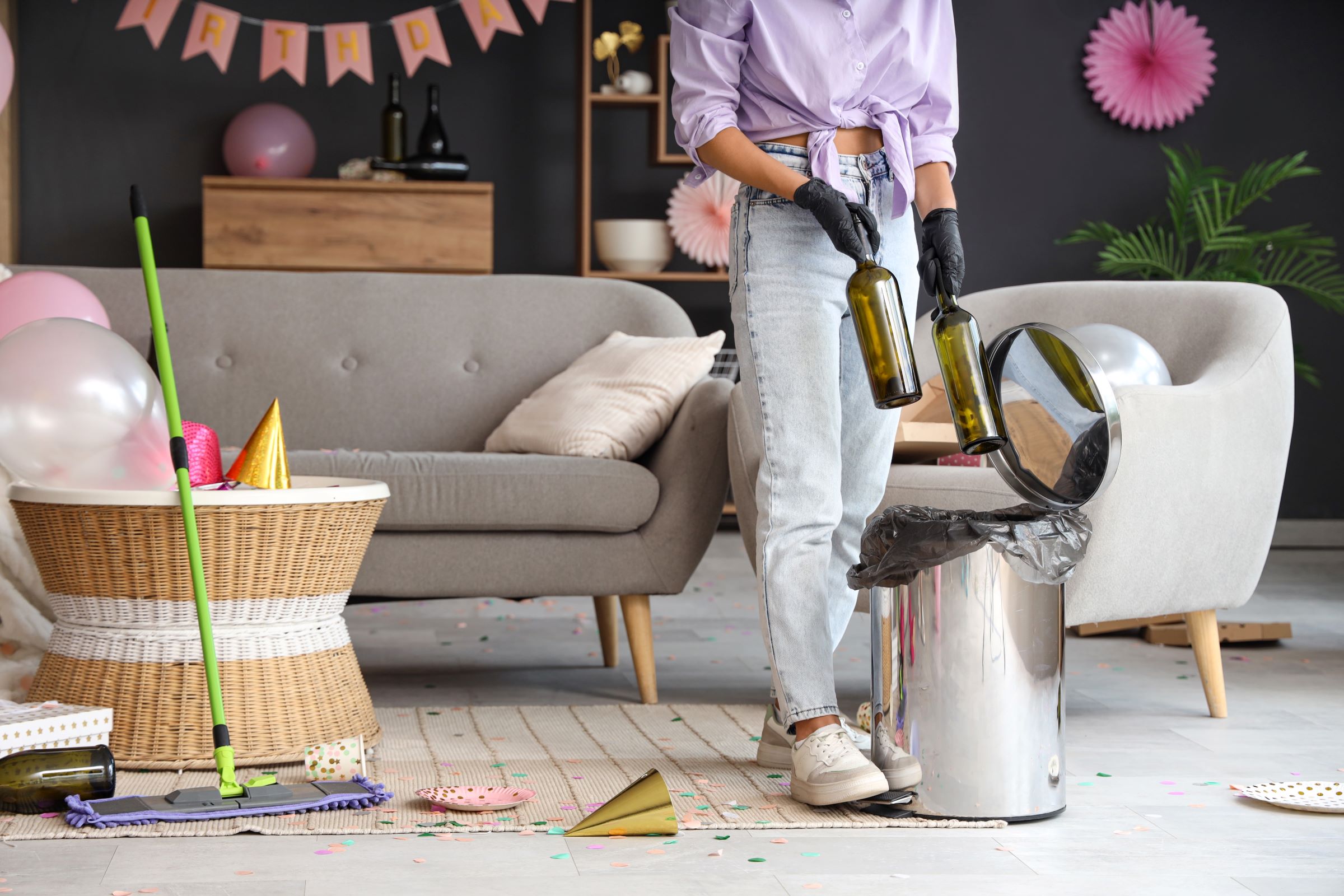 A woman throws empty wine bottles into a trash can while cleaning up after a party.