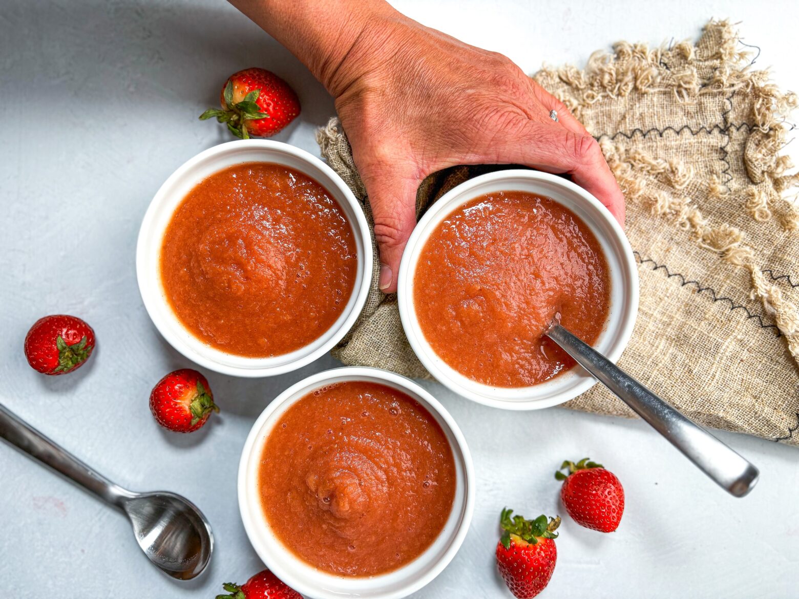 Strawberry applesauce in small dishes with a hand and whole strawberries.