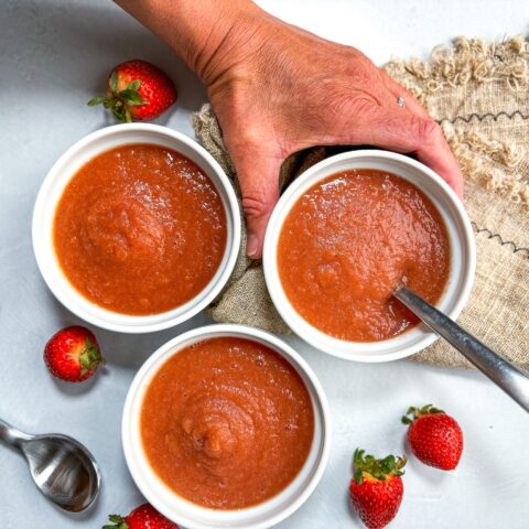 Strawberry applesauce in small dishes with a hand and whole strawberries.