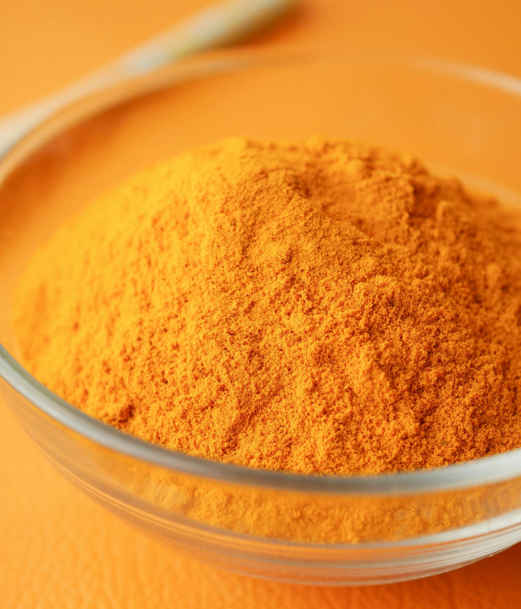 Close-up of turmeric powder in a glass bowl on an orange background.