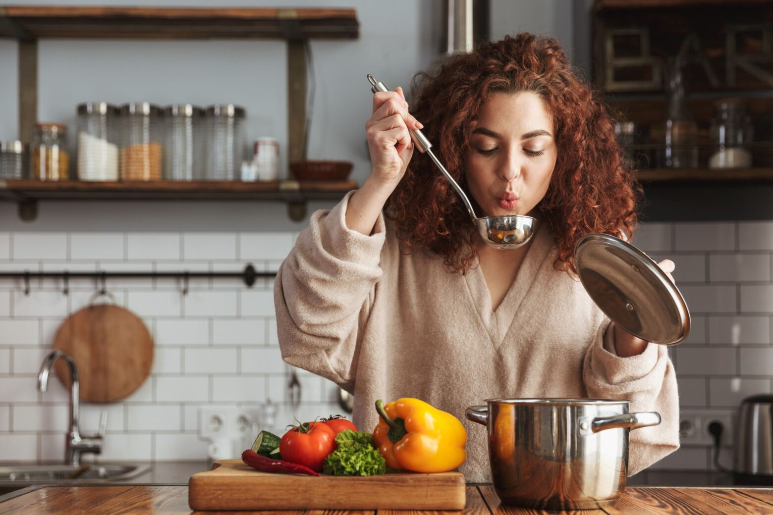 A woman stands in her kitchen, taste testing a soup made with whole vegetables.