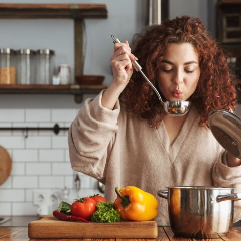 A woman stands in her kitchen, taste testing a soup made with whole vegetables.