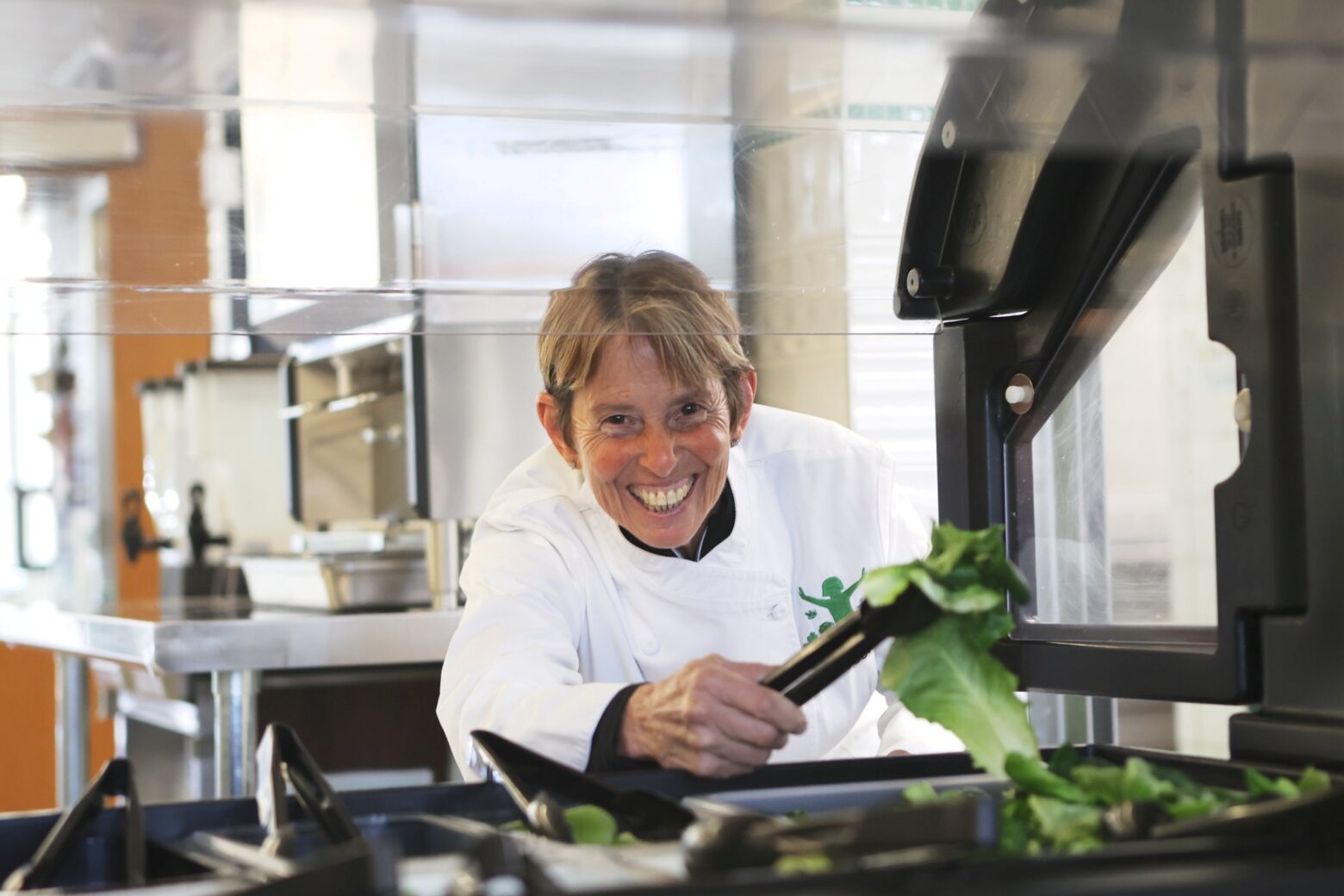 Ann Cooper holding a salad leaf in tongs as she stands behind a lunch service station.