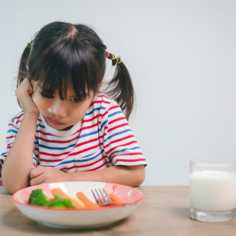 A young girl pouts over a bowl of vegetables.