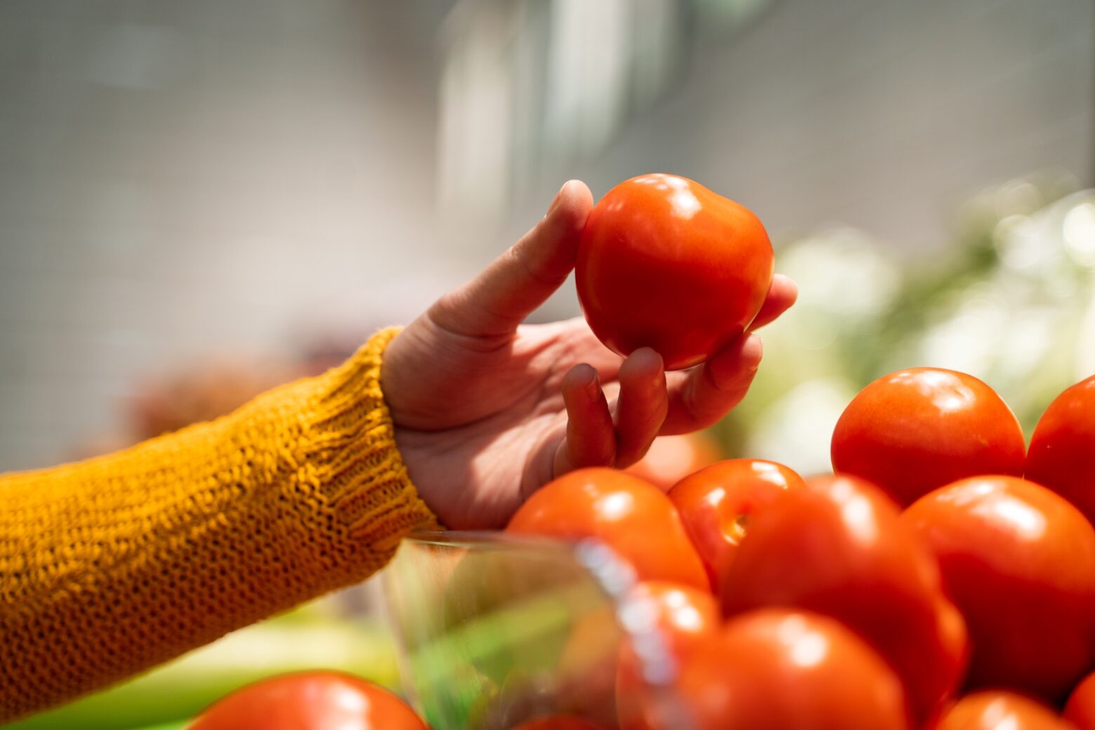 Close up of a woman holding a tomato above a pile of tomatoes in the produce section.