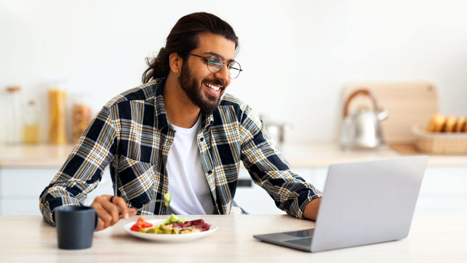 A man smiles while looking at his laptop and enjoying lunch.