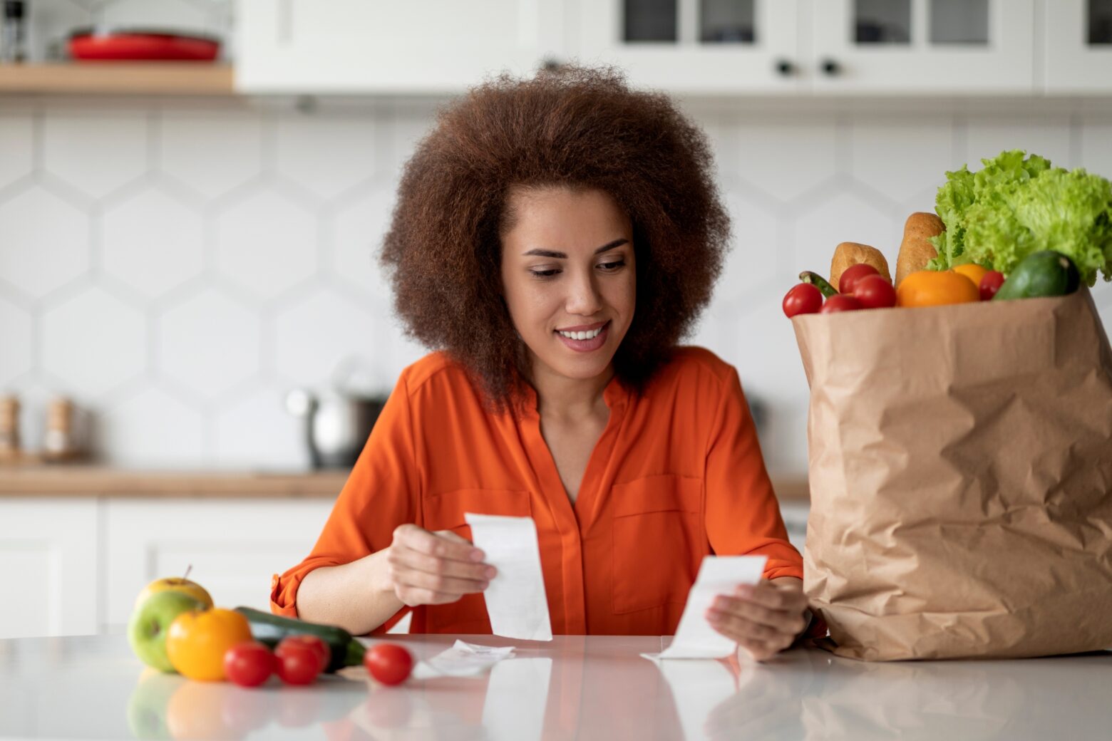 A woman examines two grocery store receipts while sitting in her kitchen