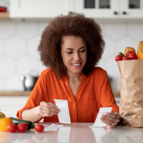 A woman examines two grocery store receipts while sitting in her kitchen