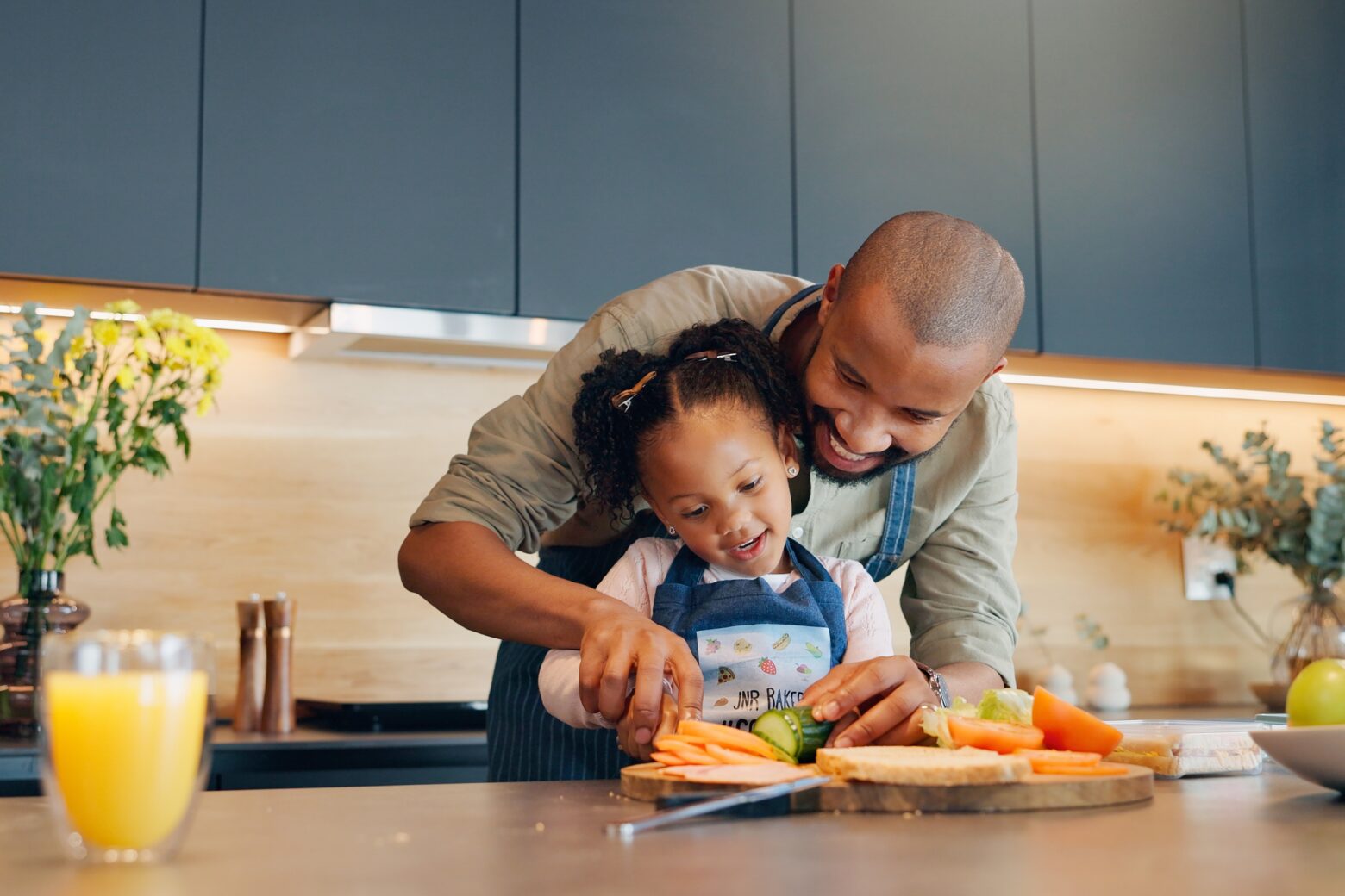 A dad helps his daughter cut vegetables in the kitchen.