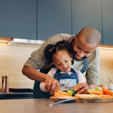 A dad helps his daughter cut vegetables in the kitchen.
