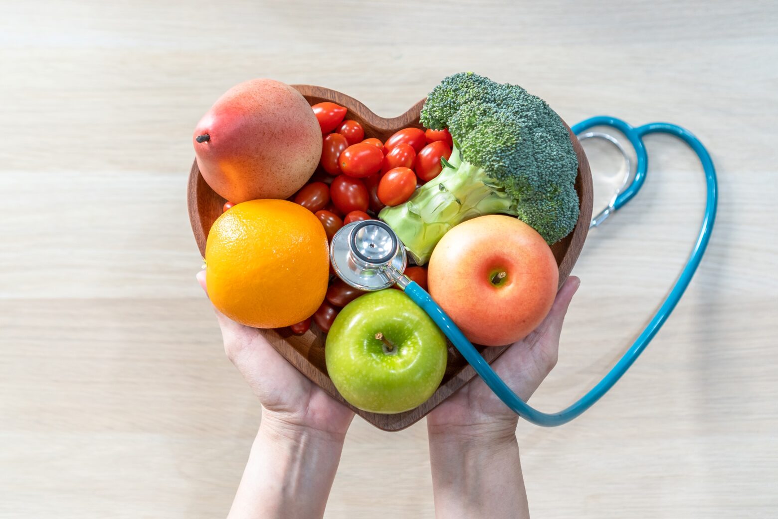 Above shot of a person holding a heart-shaped bowl filled with fruits and vegetables. A stethoscope lays across the bowl.