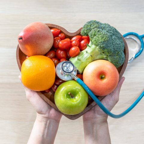 Above shot of a person holding a heart-shaped bowl filled with fruits and vegetables. A stethoscope lays across the bowl.