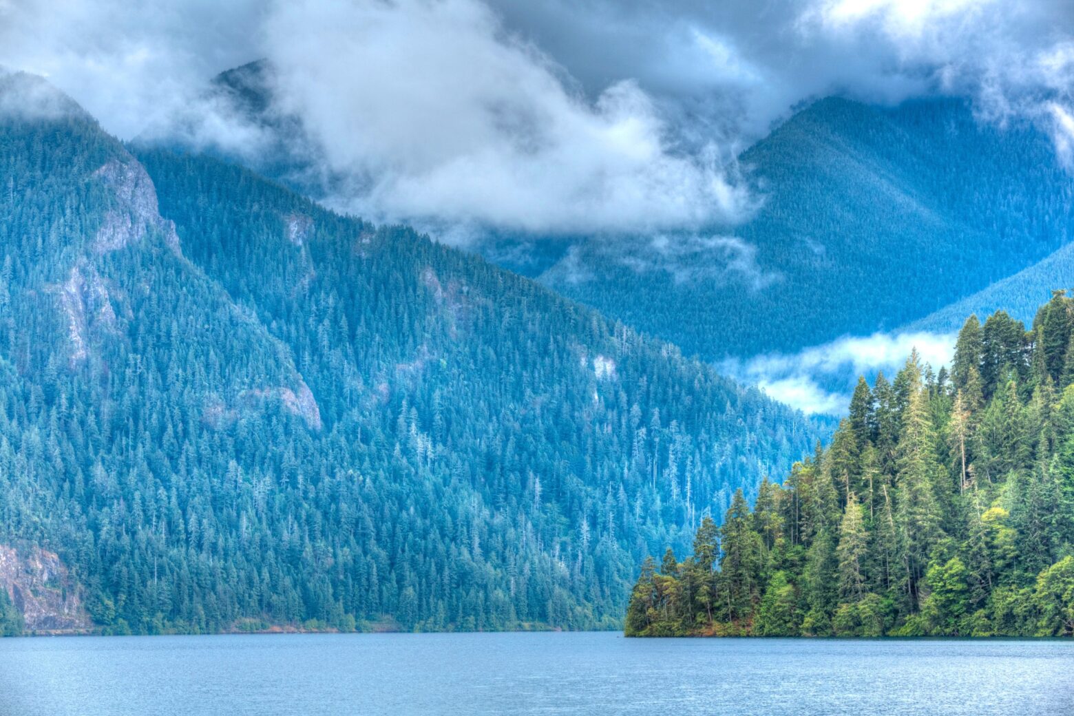 A mountain lake landscape covered with pine trees and low clouds.