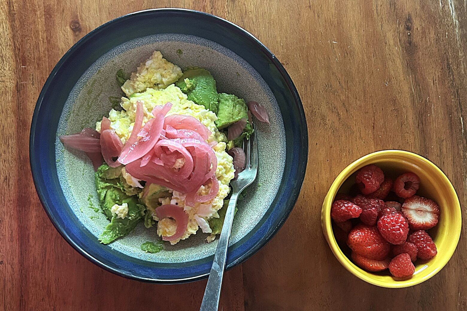 A bowl of eggs, pickled onion, and avocado next to a ramekin of berries.