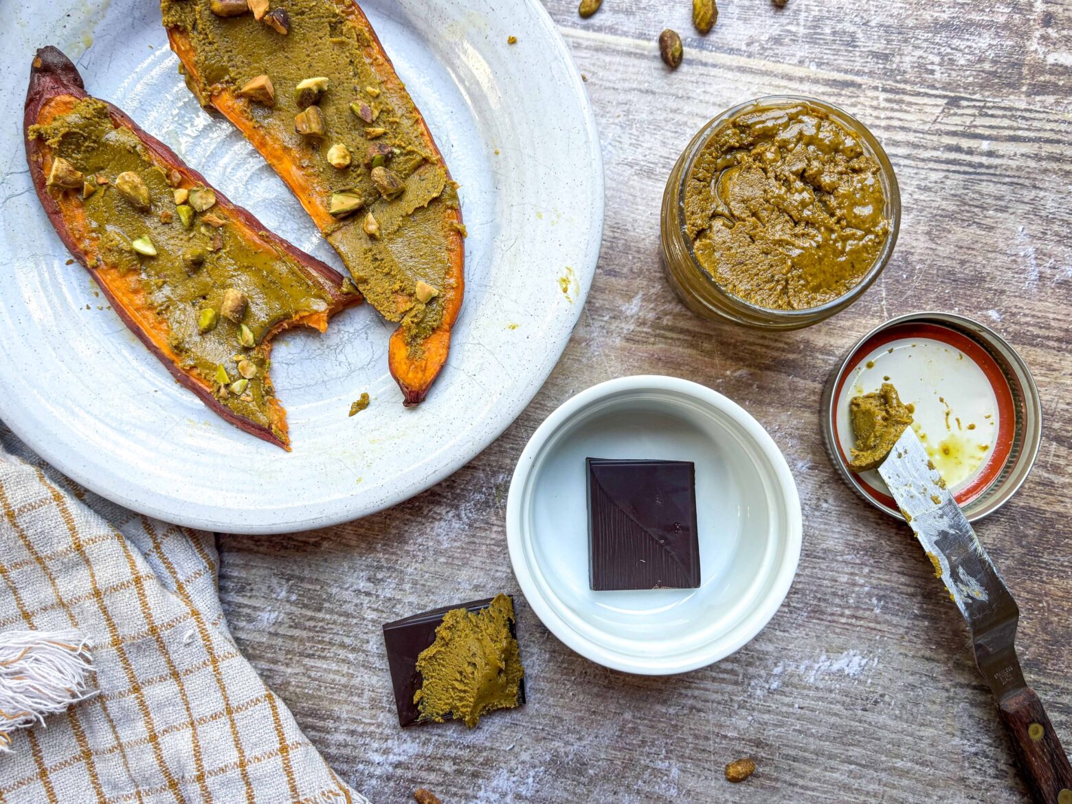 A jar of pistachio butter next to sweet potato slices and dark chocolate with pistachio butter spread over.