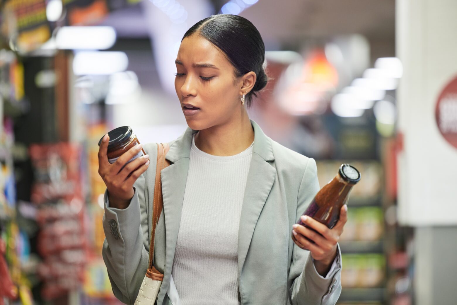 Woman holding two sauce jars in her hand at the grocery store, reading the label of one.